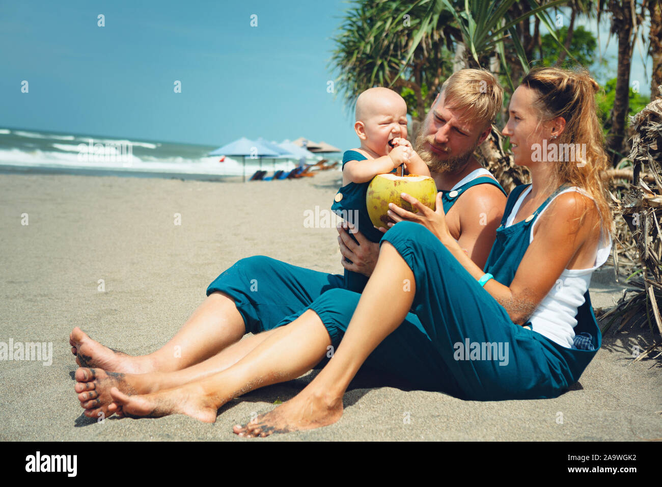 Happy hipster Familie lustig Picknick am Strand - Mutter, Vater feed baby boy. Kleiner Sohn essen sie Obst mit Spaß, trinken frische Kokosnuss. Gesunde Lebensweise. Stockfoto