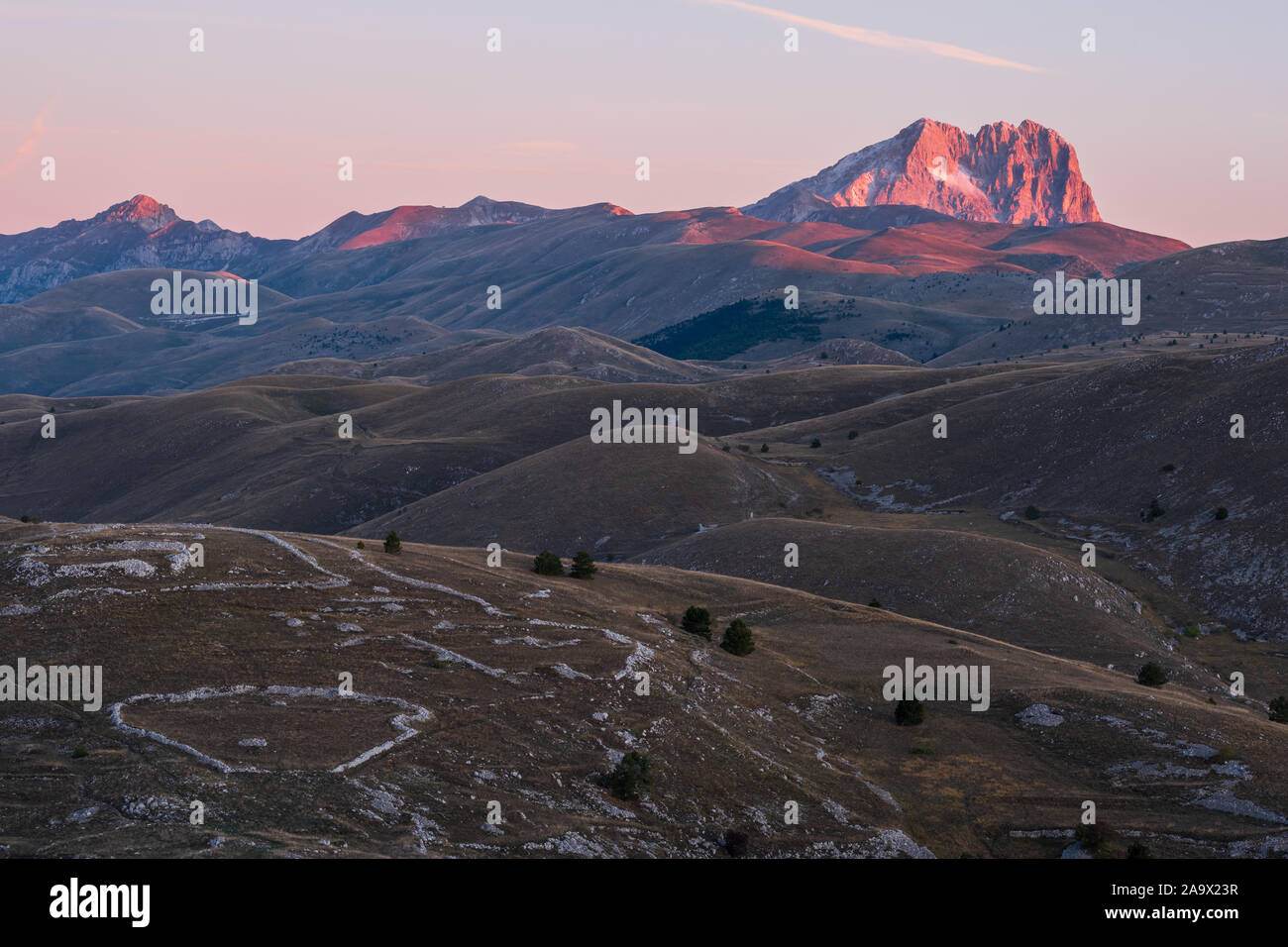 Berg Corno Grande am Horizont hinter kargen und ländliche Landschaft leuchtendem Pink im Licht des Sonnenaufgangs, Abruzzen, Italien Stockfoto