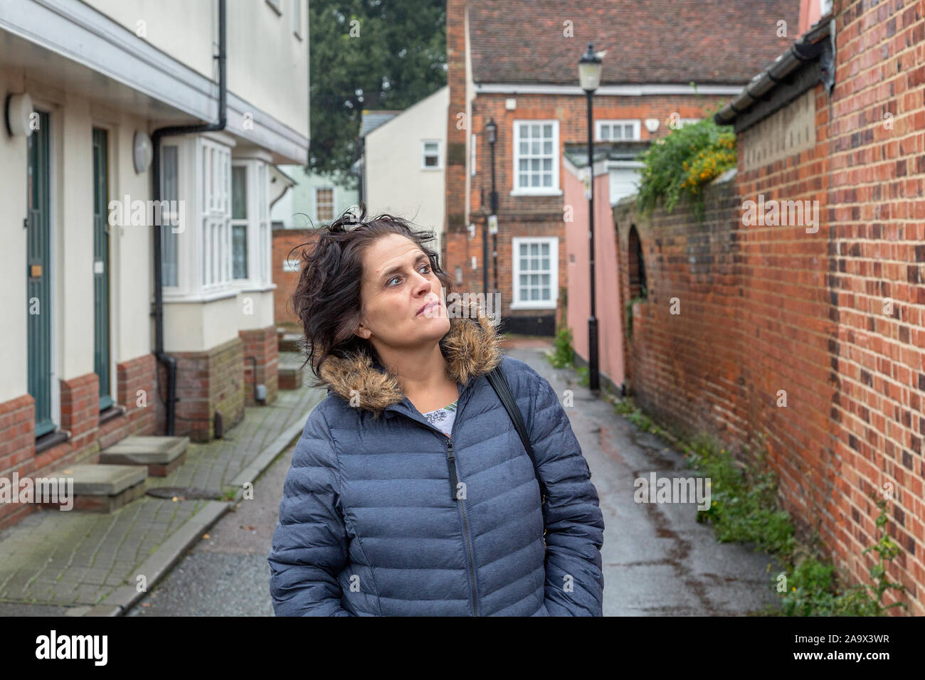 Eine Frau, die einen Regenmantel trägt, geht eine Straße entlang. Der Boden und ihre Haare sind vom Regen nass. Stockfoto