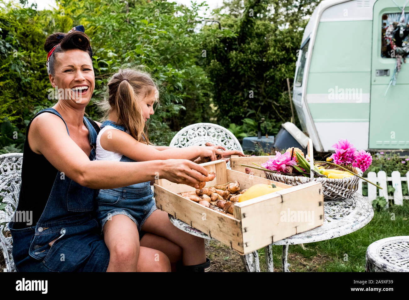 Lachende Frau und Mädchen am Tisch sitzen in einem Garten, Holzkiste mit Gemüse und Korb mit Dahlien. Stockfoto