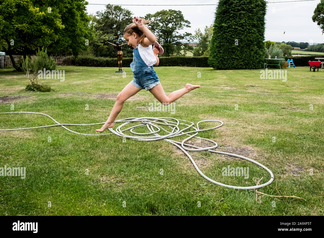 Mädchen tragen Jeans Latzhose über einen Gartenschlauch auf einem Rasen springen. Stockfoto