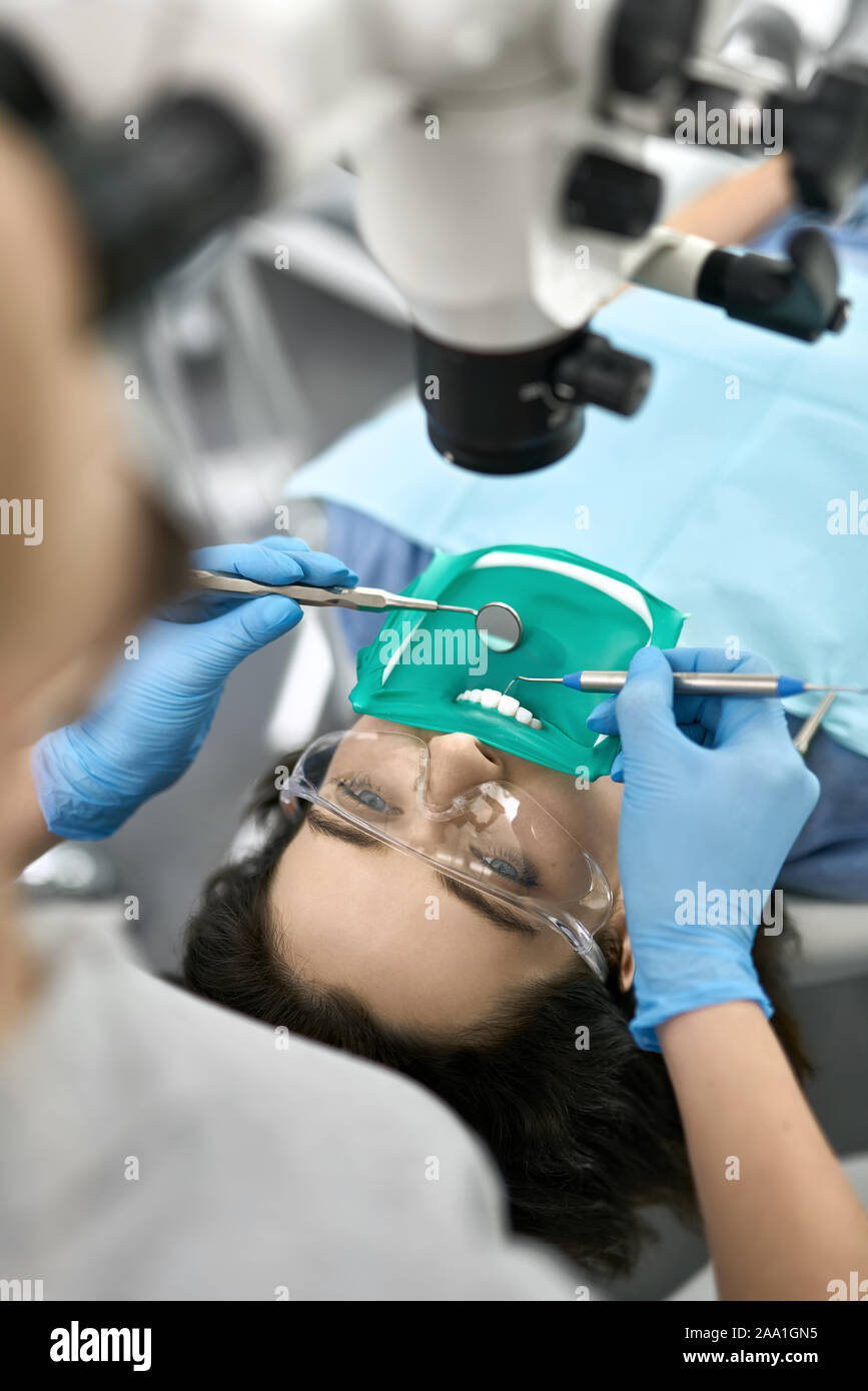 Nettes Mädchen in eine Schutzbrille und eine Wange Gurtaufroller mit einem grünen Kofferdam in einer Zahnarztpraxis. Zahnarzt in blau Latex Handschuhe untersucht ihre Zähne mit Stockfoto