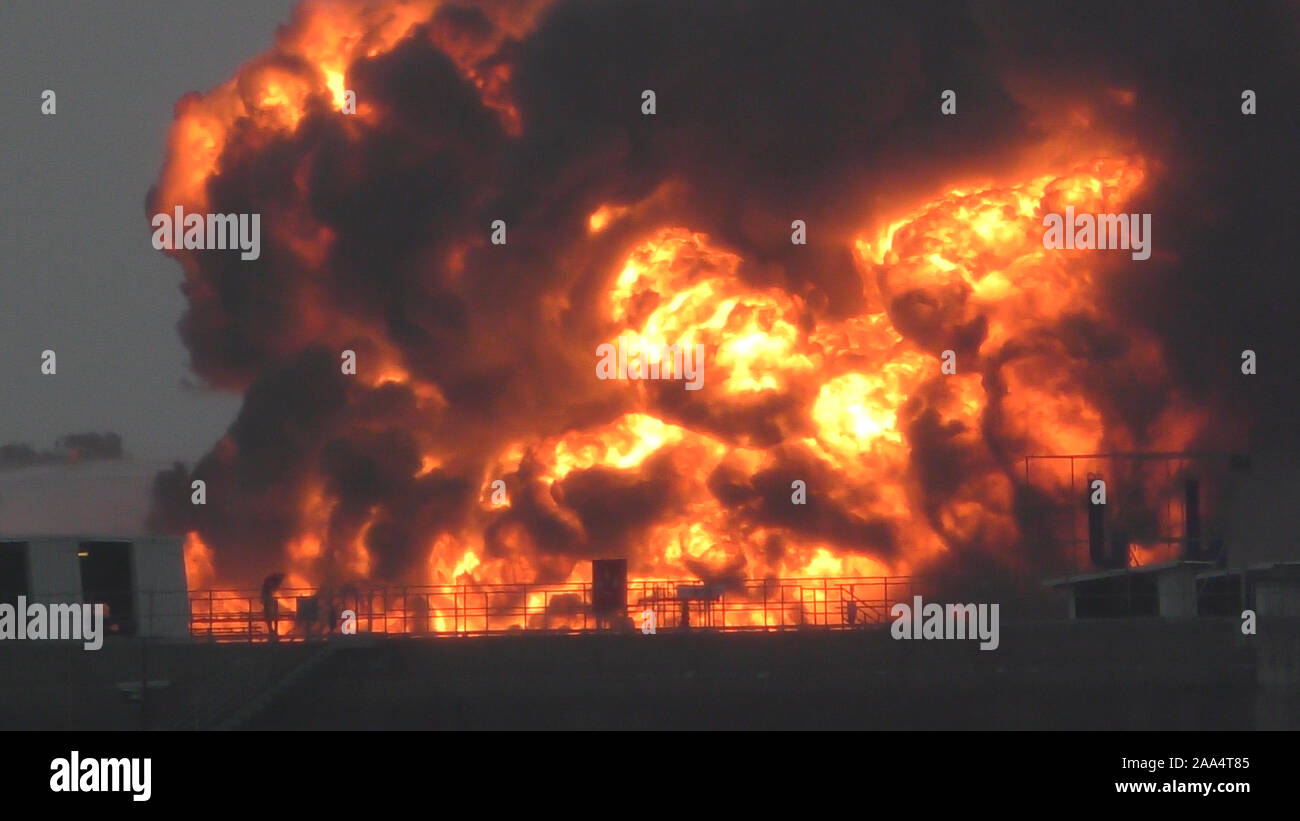 Riesige Feuer Flamme bei der Verbrennung von Öl behälter in der Raffinerie. Feuerwehrmann Wasserstrahl löschen das Feuer begann in der Nähe einer Tankstelle Lagertank. Schwarzer Regen, saurer Regen, Smog, Rauch, Asche, Umweltverschmutzung, verschwommenes Luft Stockfoto