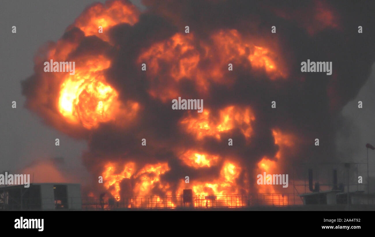 Riesige Feuer Flamme bei der Verbrennung von Öl behälter in der Raffinerie. Feuerwehrmann Wasserstrahl löschen das Feuer begann in der Nähe einer Tankstelle Lagertank. Schwarzer Regen, saurer Regen, Smog, Rauch, Asche, Umweltverschmutzung, verschwommenes Luft Stockfoto