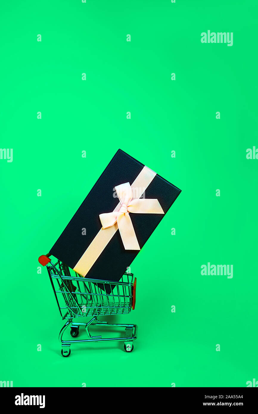 Urlaub Geschenk, Schwarz Grün Freitag Konzept mit Geschenkbox und rosa Schleife in einem Metall Einkaufswagen, Korb auf grüne Farbe Hintergrund. Shopping. Outfit. Flach, Ansicht von oben, Platz für Text. Stockfoto