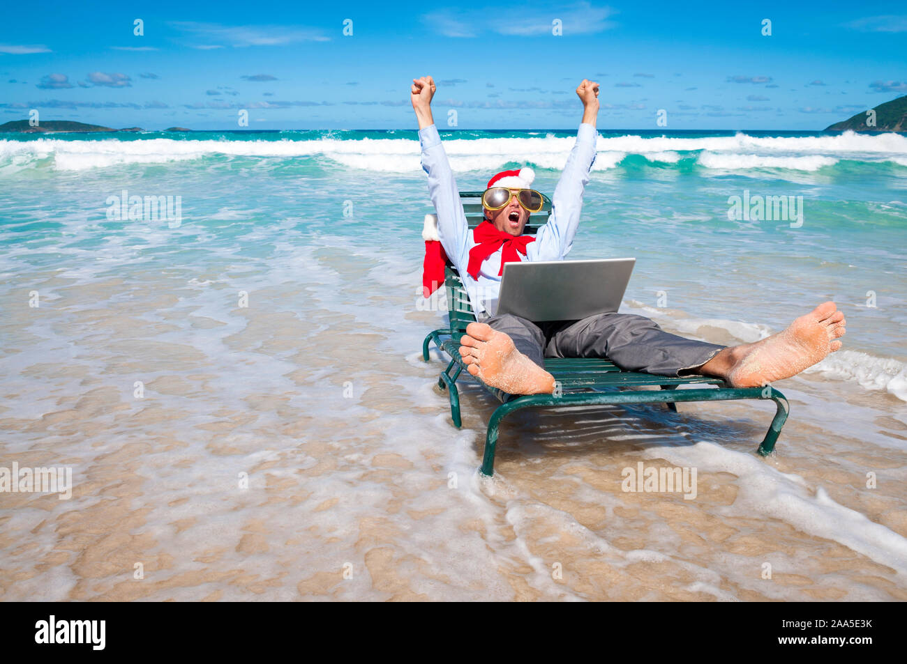 Aufgeregt Geschäftsmann in Santa Hut, Sonnenbrille und einem grossen roten Bogen Weihnachten feiern mit seinem Laptop an einem tropischen Strand Stockfoto