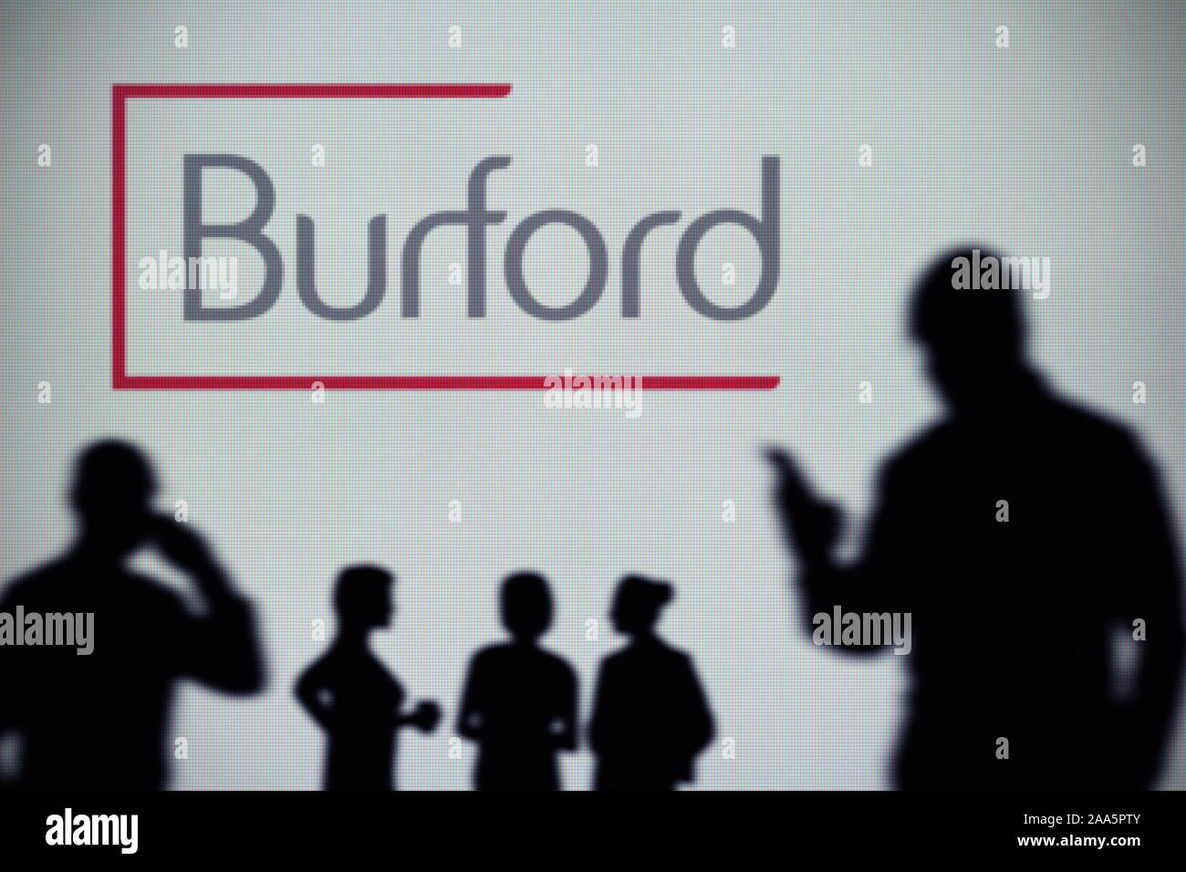 Die burford Hauptstadt Logo ist auf einen LED-Bildschirm im Hintergrund, während eine Silhouette Person ein Smartphone verwendet (nur redaktionelle Nutzung) Stockfoto