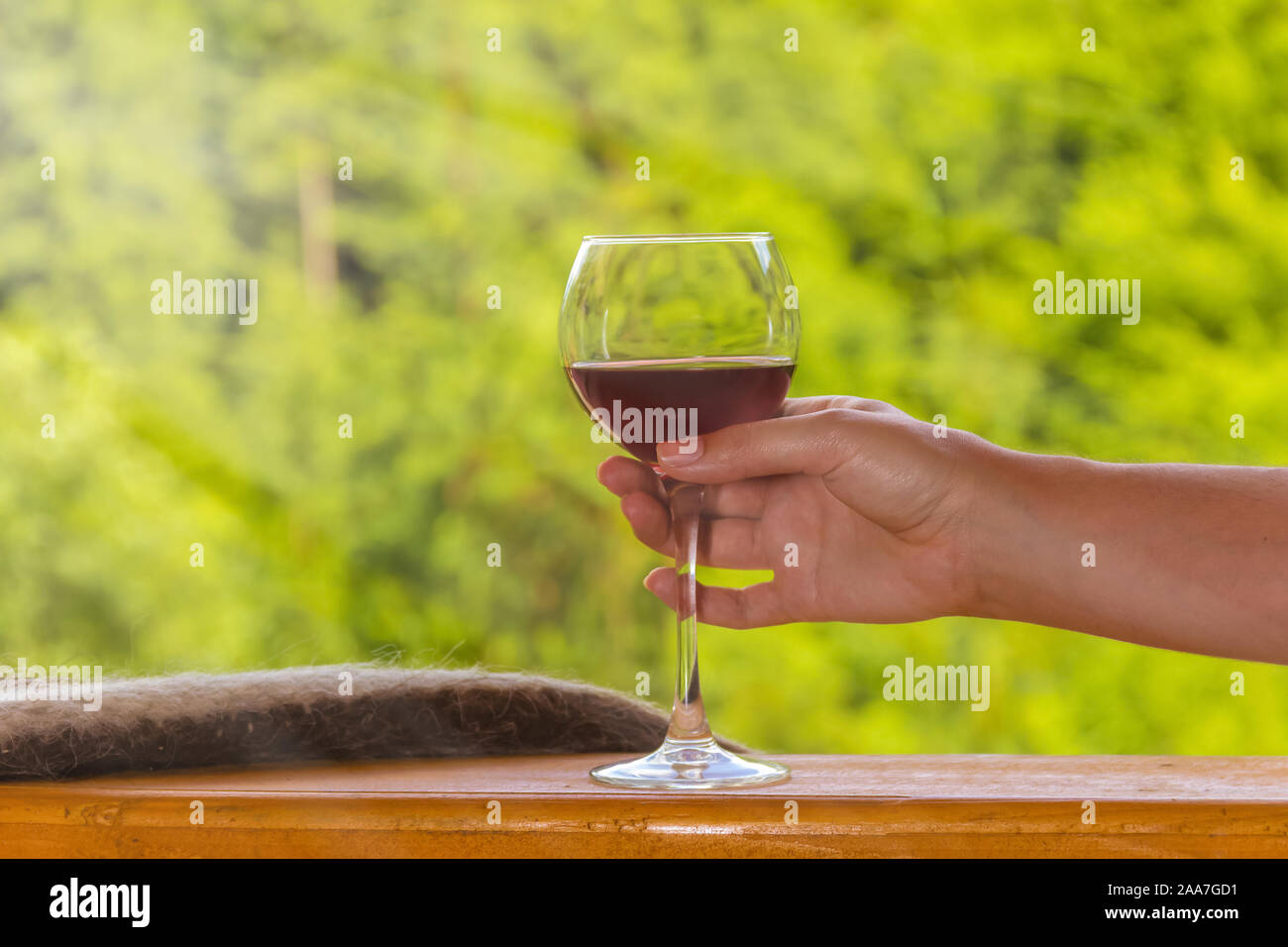 Ein Glas Rotwein in eine weibliche Hand auf einem Holzdeck Landhaus auf einem Hintergrund von grünen Wald. Sommer Urlaub Konzept Stockfoto