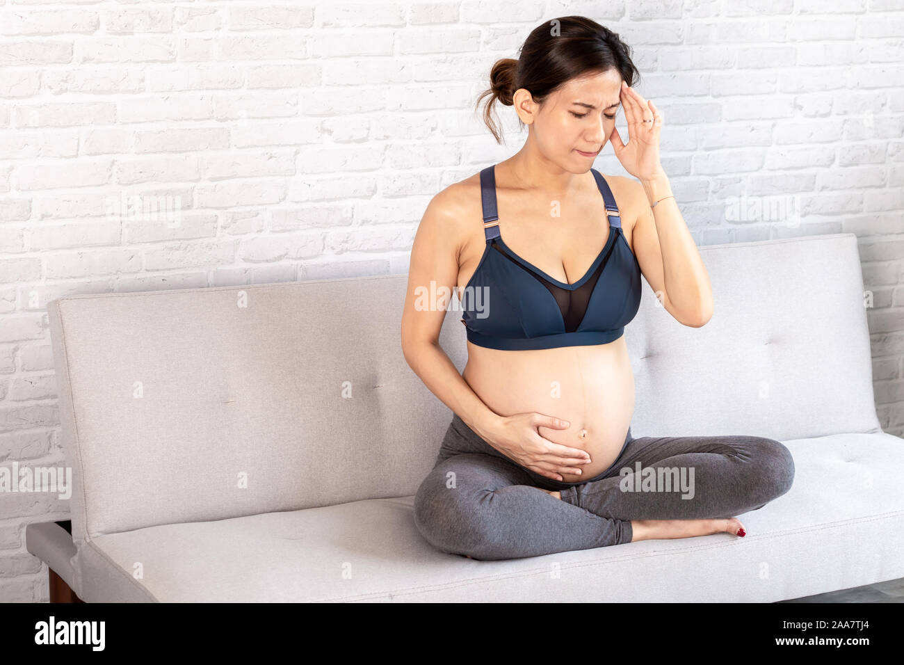 Müde Asiatischen schwangere Frau berühren Kopf und Magen leiden unter Kopfschmerzen auf dem Sofa sitzen mit gekreuzten Beinen Stockfoto