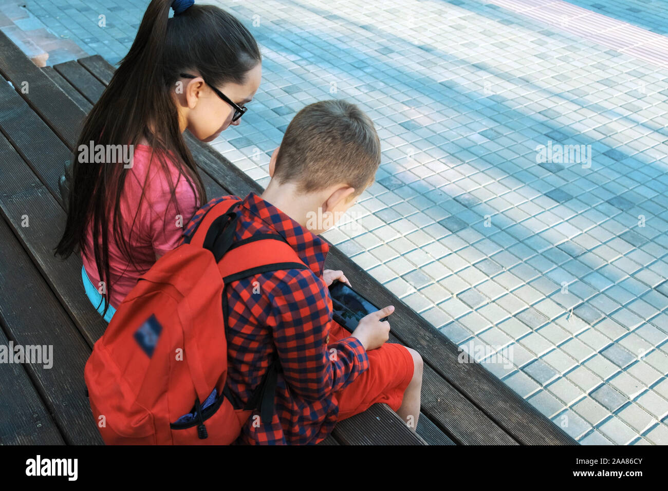 Die ältere Schwester und Bruder sitzen auf der Straße, an Smartphones. Kinder gehen nach der Schule Stockfoto