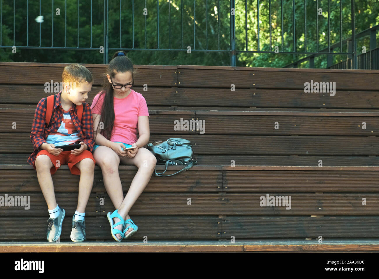 Die ältere Schwester und Bruder sitzen auf der Straße, an Smartphones. Kinder gehen nach der Schule Stockfoto