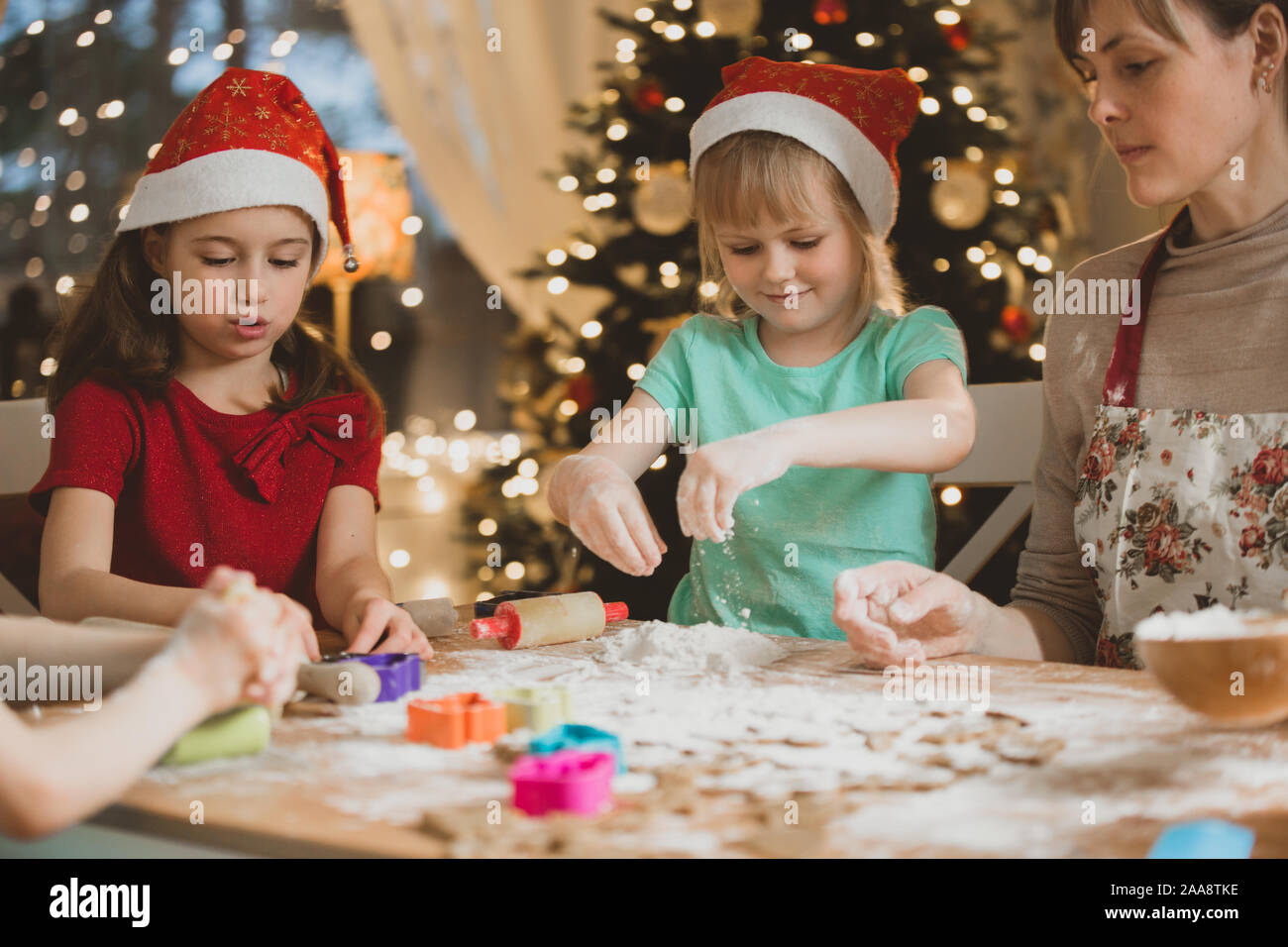 Mutter und Kinder in roten Hüte kochen Lebkuchen cookies und Spielen. Schönes Wohnzimmer mit Lichtern und Weihnachtsbaum, Tisch mit Laterne. Stockfoto