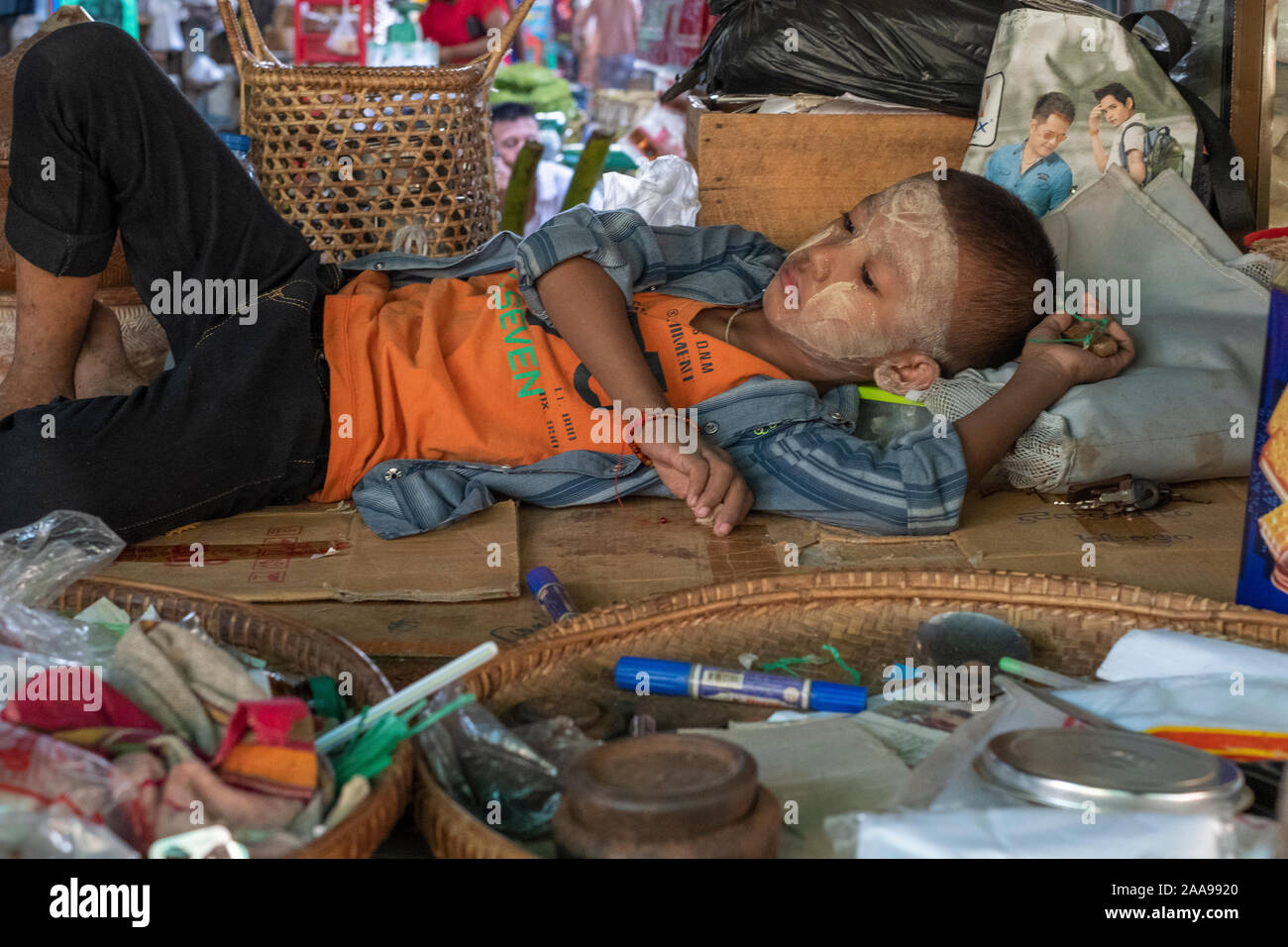 Die jungen burmesischen Jungen 6-8 Jahre alt mit einem pulverförmigen Gesicht (Sonnenschutz) liegen auf einer Decke im Markt. seine Mutter als das Leben geht Abschaltdruck durch Stockfoto