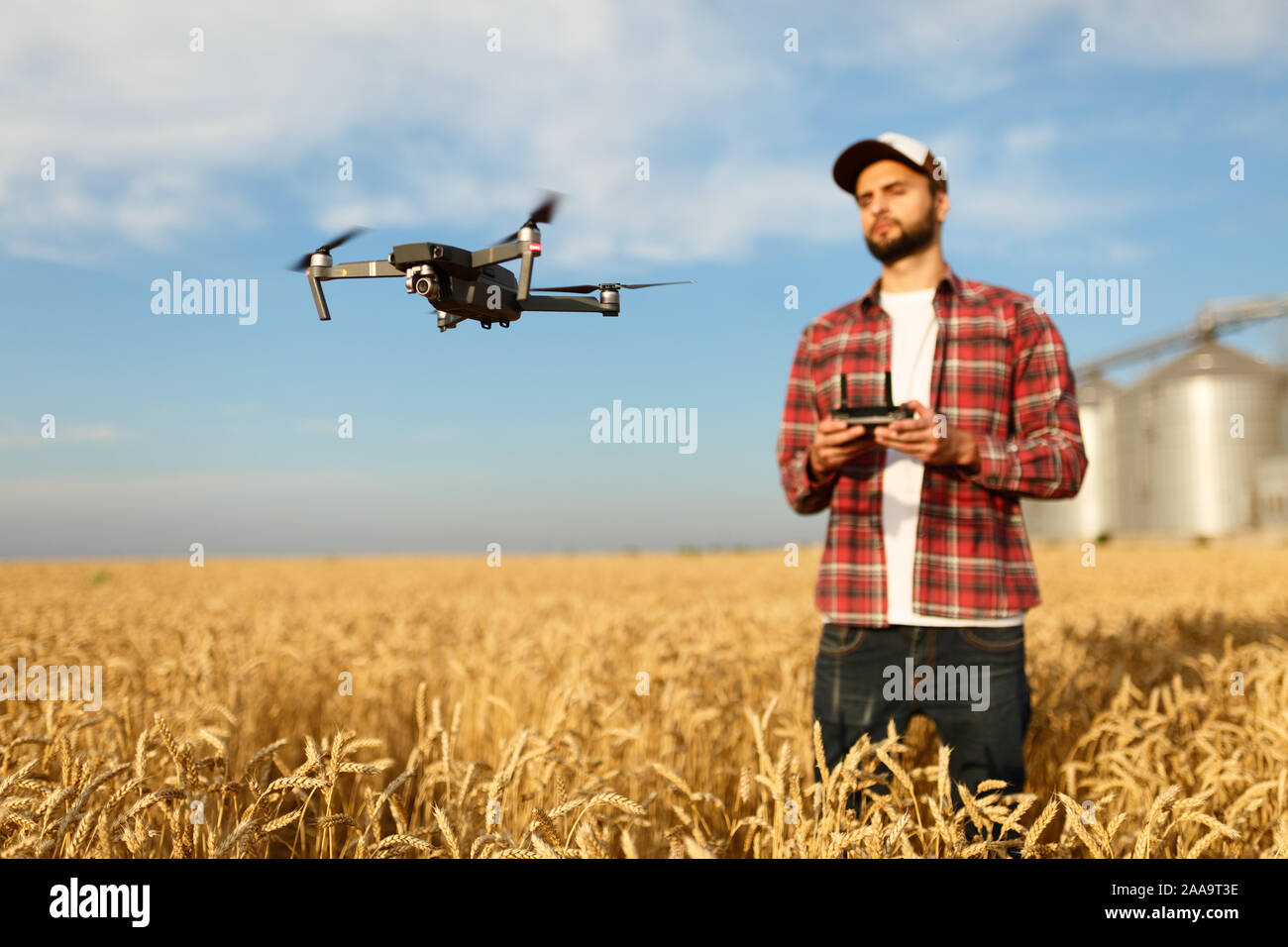 Drone schwebt vor Bauer mit Fernbedienung in der Hand in der Nähe des Kornelevators. Quadcopter fliegt in der Nähe von Pilot. Agrarwissenschaftler, Luftaufnahmen und Stockfoto