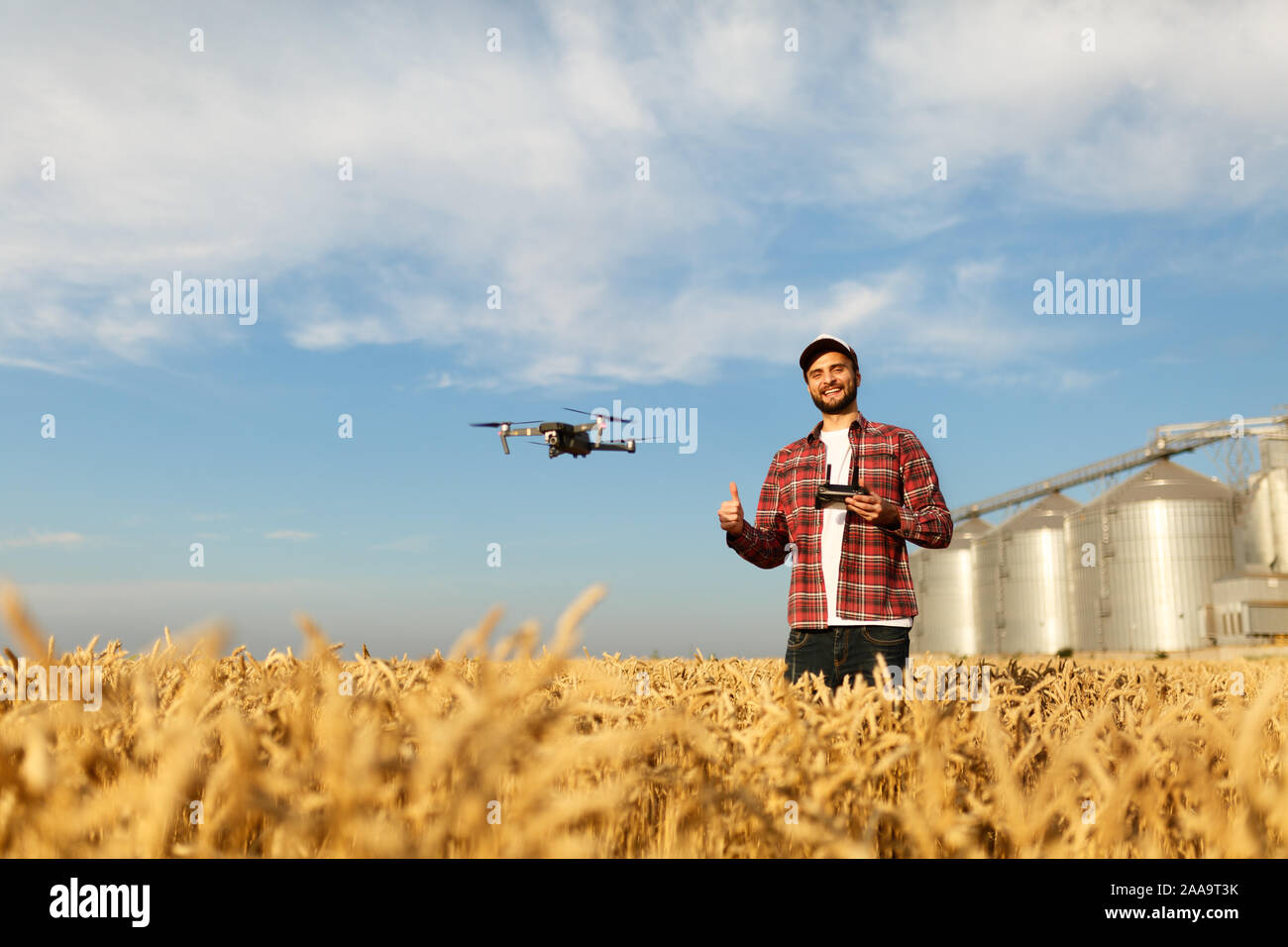 Drone schwebt vor Bauer mit Fernbedienung in der Hand in der Nähe des Kornelevators. Quadcopter fliegt in der Nähe von Pilot. Agrarwissenschaftler, Luftaufnahmen und Stockfoto