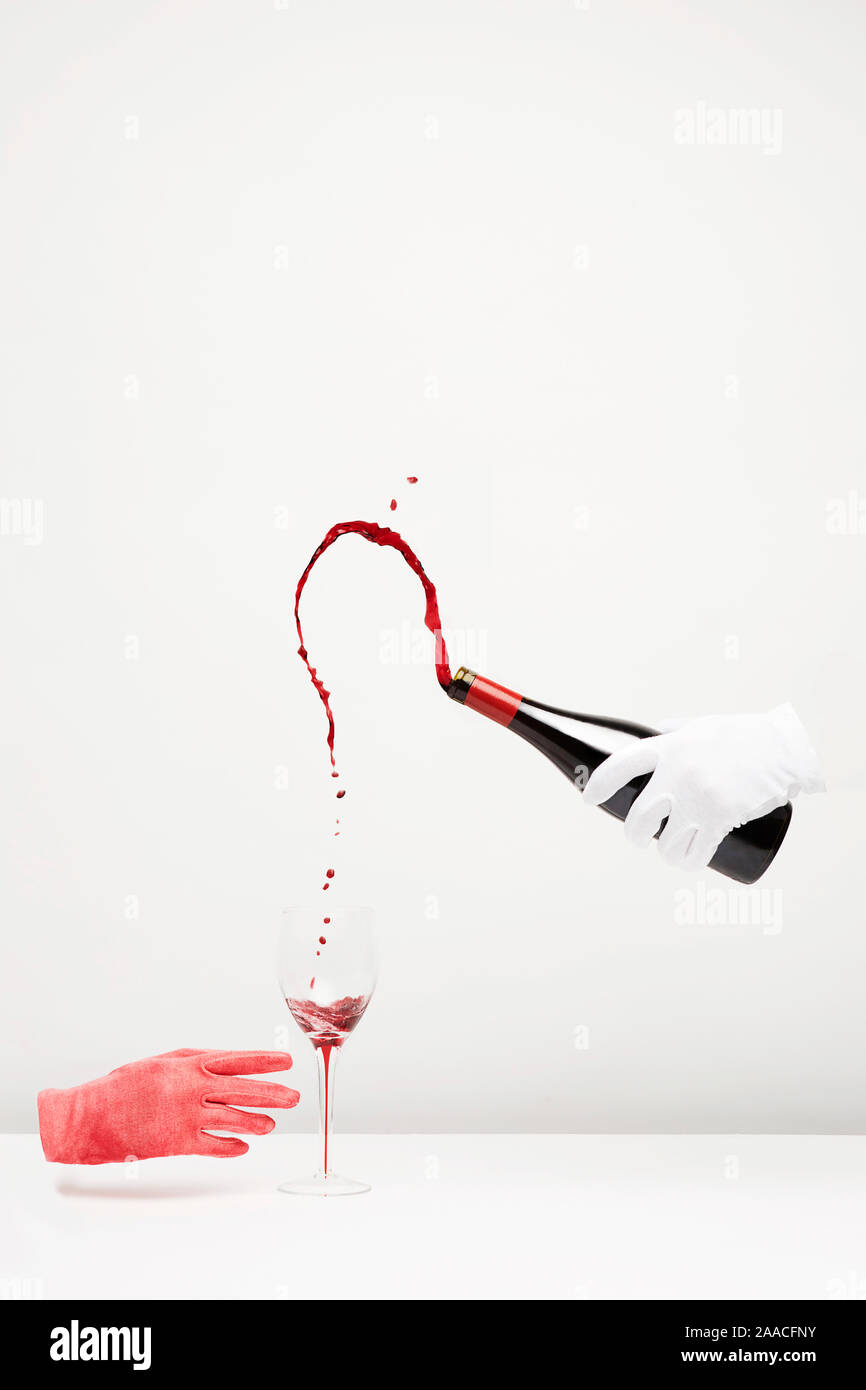 Surrealistische Bild von 2 Hände in Handschuhen auf weißem Hintergrund und ein Glas Wein auf einem weißen Tisch. Einerseits hält eine Flasche Wein. Stockfoto