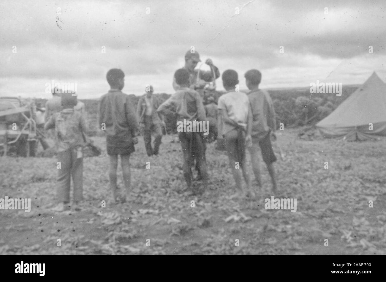 Eine kleine Gruppe von Kindern, von der Rückseite, stand in einem landwirtschaftlichen Gebiet an einem sonnigen Tag, einen uniformierten Offizier ein Objekt manipulieren, mit Zelten und anderen Soldaten (darunter ein Mann auf Krücken) im Hintergrund, Vietnam, 1965. () Stockfoto