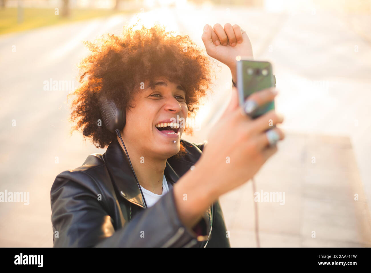 Hübscher junger Mann in Schwarz mit Afro Frisur hören Musik mit mobilen und Kopfhörer in einer sonnigen Stadt Stockfoto