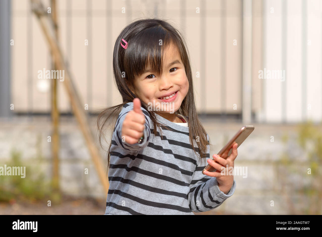Cute glückliche junge Mädchen mit Grübchen lächelnd in die Kamera, als Sie einen Daumen nach oben Geste mit der Hand gibt, während Sie ein Handy im Freien in sunsh Stockfoto