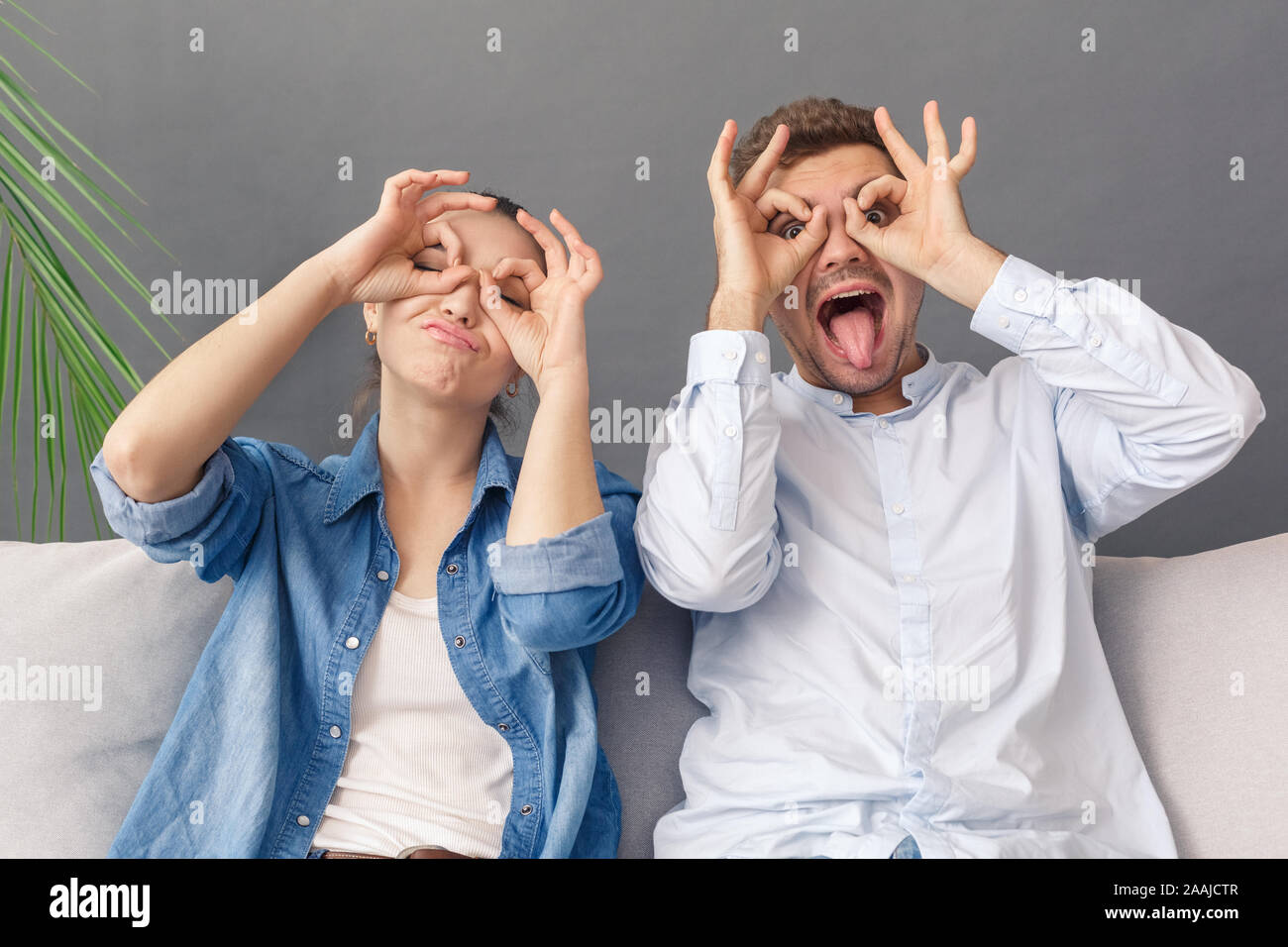 Relationnship Konzept. Junges Paar sitzt auf einem Sofa studio isoliert auf grau, Brillen mit Händen Grimassen verspielt Stockfoto