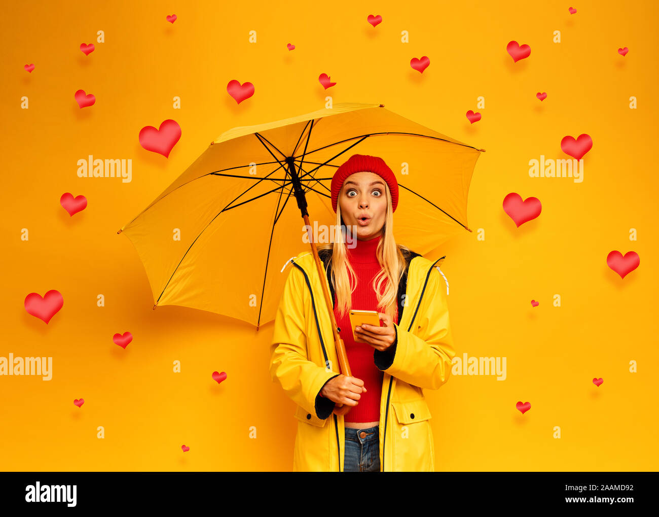 Blonde süße Mädchen sich schützt mit Schirm durch den Regen der Herzen auf Ihrem Smartphone. glücklich und überrascht Ausdruck Gesicht. Hintergrund Stockfoto