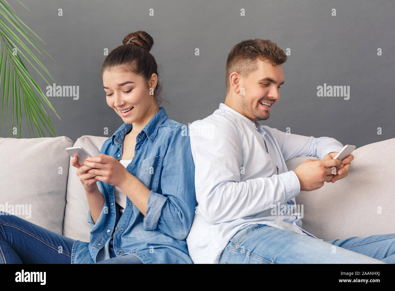 Moderne Relatonship. Junges Paar sitzt auf einem Sofa studio isoliert auf Grau online plaudern auf dem Smartphone lächelnd Frohe Stockfoto