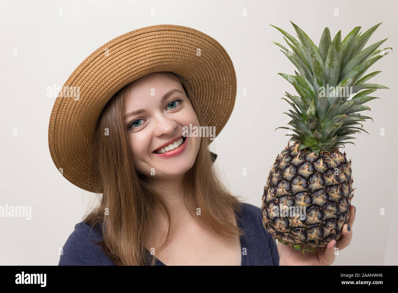 Portraitfoto eines hübschen Mädchens in dunklem Hemd und Hut mit einer ganzen Ananas. Weißer Hintergrund, warme Farben Stockfoto