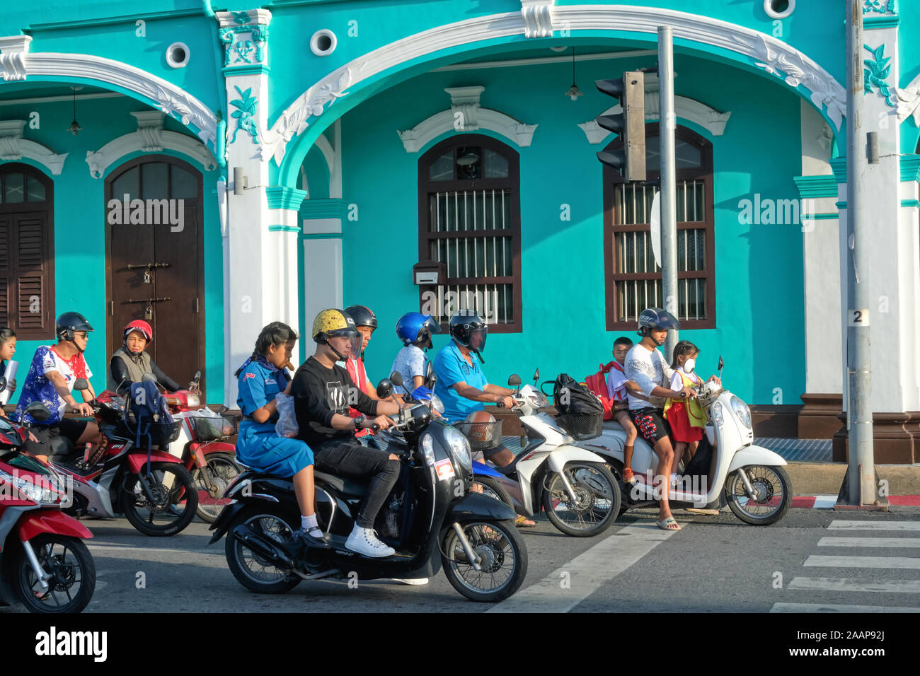 Motorradfahrer, einige mit ihren Kindern auf ihren Fahrrädern, an einer roten Ampel anhalten vor einem chinesisch-portugiesischen Stil Haus in der Stadt Phuket, Thailand Stockfoto
