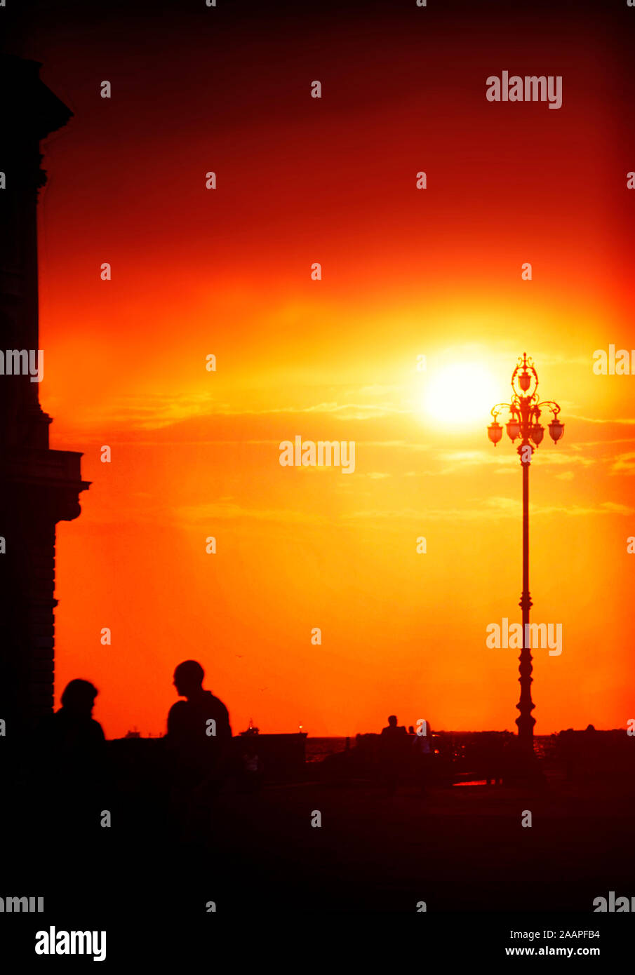 Roter Himmel bei Sonnenuntergang über dem Meer promenade und unita' d'italia Square in Triest, Italien mit Silhouetten von einem Gebäude und Menschen im Licht Stockfoto