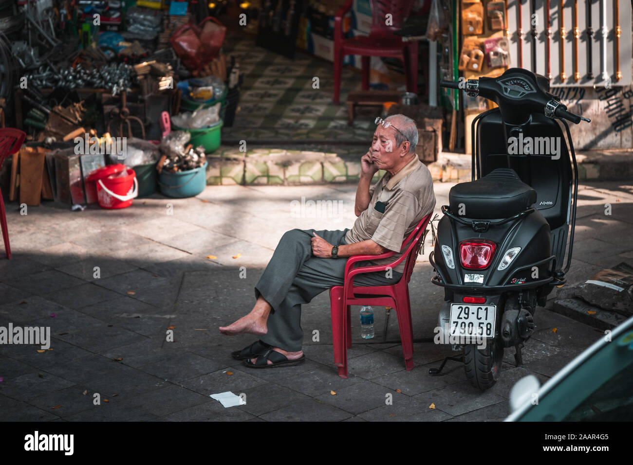 Hanoi, Vietnam - 12. Oktober 2019: Eine alte ältere Menschen asiatischer Mann sitzt auf einem Stuhl aus Kunststoff neben einem Moped auf dem Telefon Stockfoto