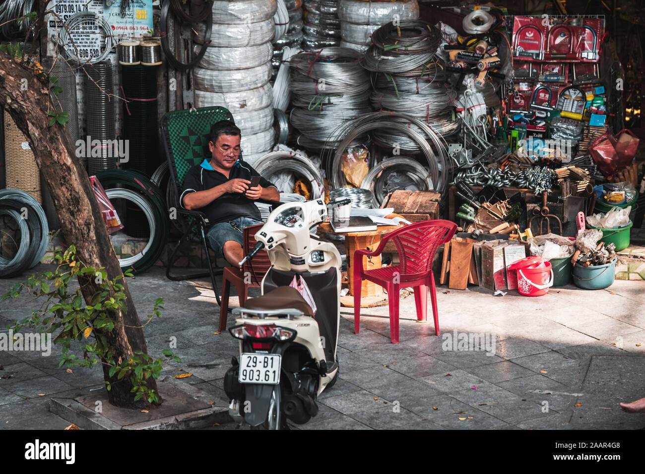 Hanoi, Vietnam - 12. Oktober 2019: Ein asiatischer Mann sitzt auf einem Stuhl neben einem Moped vermeiden der heißen Hitze des Tages texting stürzte Stockfoto