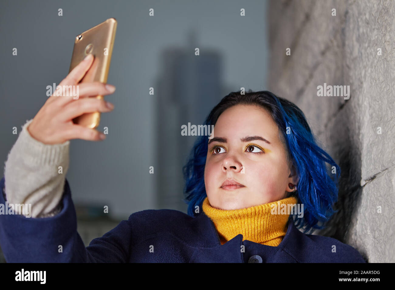 Eine junge Frau mit blau gefärbten Haaren und einem Mantel der gleichen Farbe Verwendet ein Smartphone ein selfie zu nehmen, oder ein Video im Freien am Abend zu schießen. Ein chubb Stockfoto