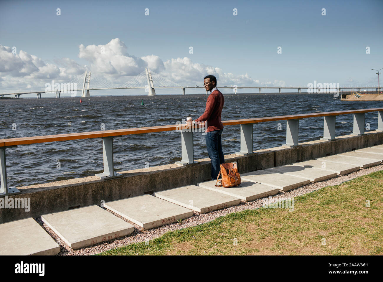 Junge Mann sitzt auf Geländer am Meer, Hamburger essen, trinken Kaffee Stockfoto