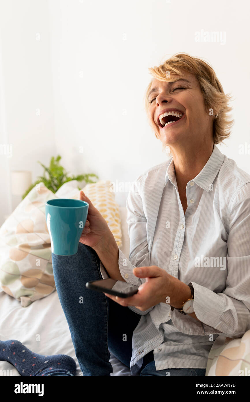 Lachen, reife Frau auf dem Bett sitzen zu Hause holding Smartphone und Kaffee Tasse Stockfoto