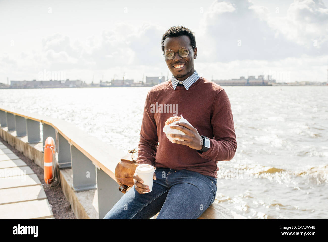 Junge Mann sitzt auf Geländer am Meer, Hamburger essen, trinken Kaffee Stockfoto