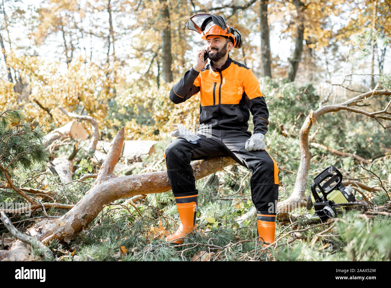 Professionelle lumberman in schützende Arbeitskleidung am Telefon sprechen, beim Sitzen auf der gefällten Baumes, ruht nach der harten Arbeit im Wald Stockfoto