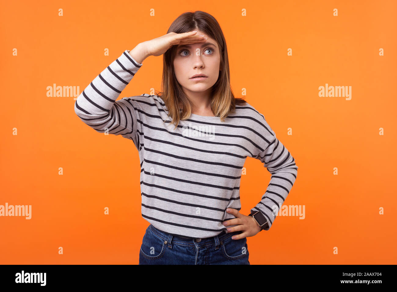 Portrait von Konzentriertem neugierige junge Frau mit braunen Haaren in Long Sleeve Striped Shirt ständigen Suchen weit weg mit der Hand über die Augen, erstaunt anstarren Stockfoto