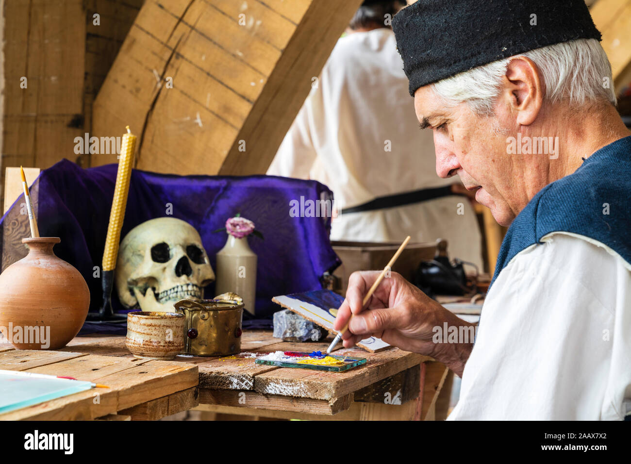 Mittelalterliche Geschichte. Nahaufnahme, reifer Mann im mittelalterlichen Kostüm während Bank sitzen und Malen ein Schädel auf dunkelblauem Hintergrund. Stockfoto