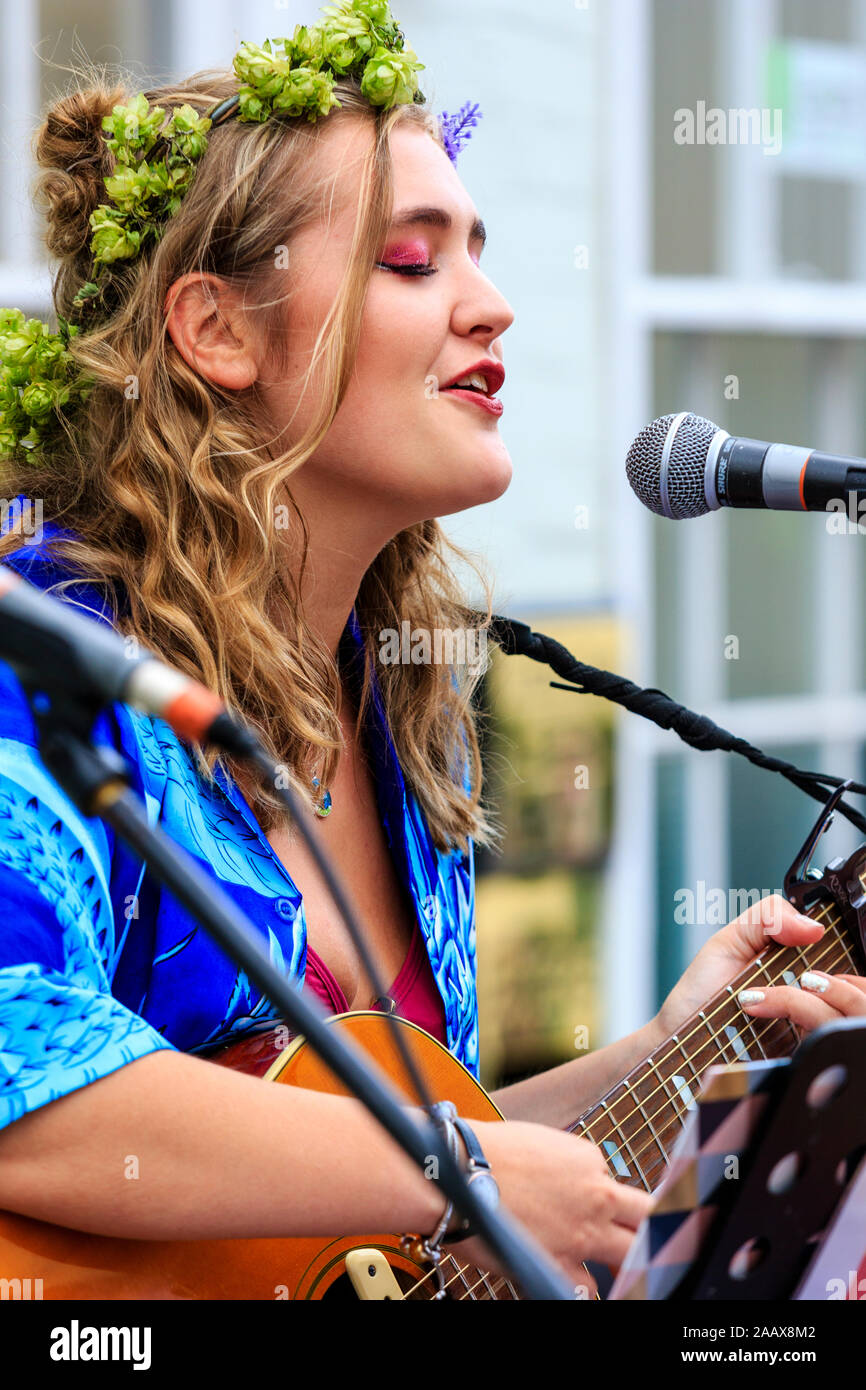 Maisy Beth, junge blonde Volkssänger, Gesang während der Wiedergabe akustischer Gitarre in die Straße an der Faversham Hop Festival. Trägt Girlande von Hopfen. Stockfoto