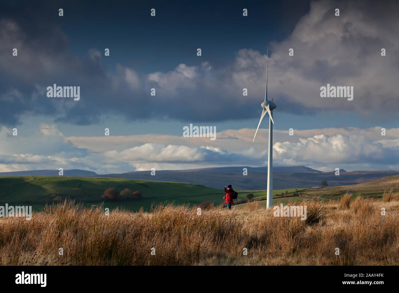 Walker auf dem Hügelkamm, der von Windturbinen mit dramatischem Himmel am späten Nachmittag in den South Wales Valleys in den Schatten gestellt wurde. Stockfoto
