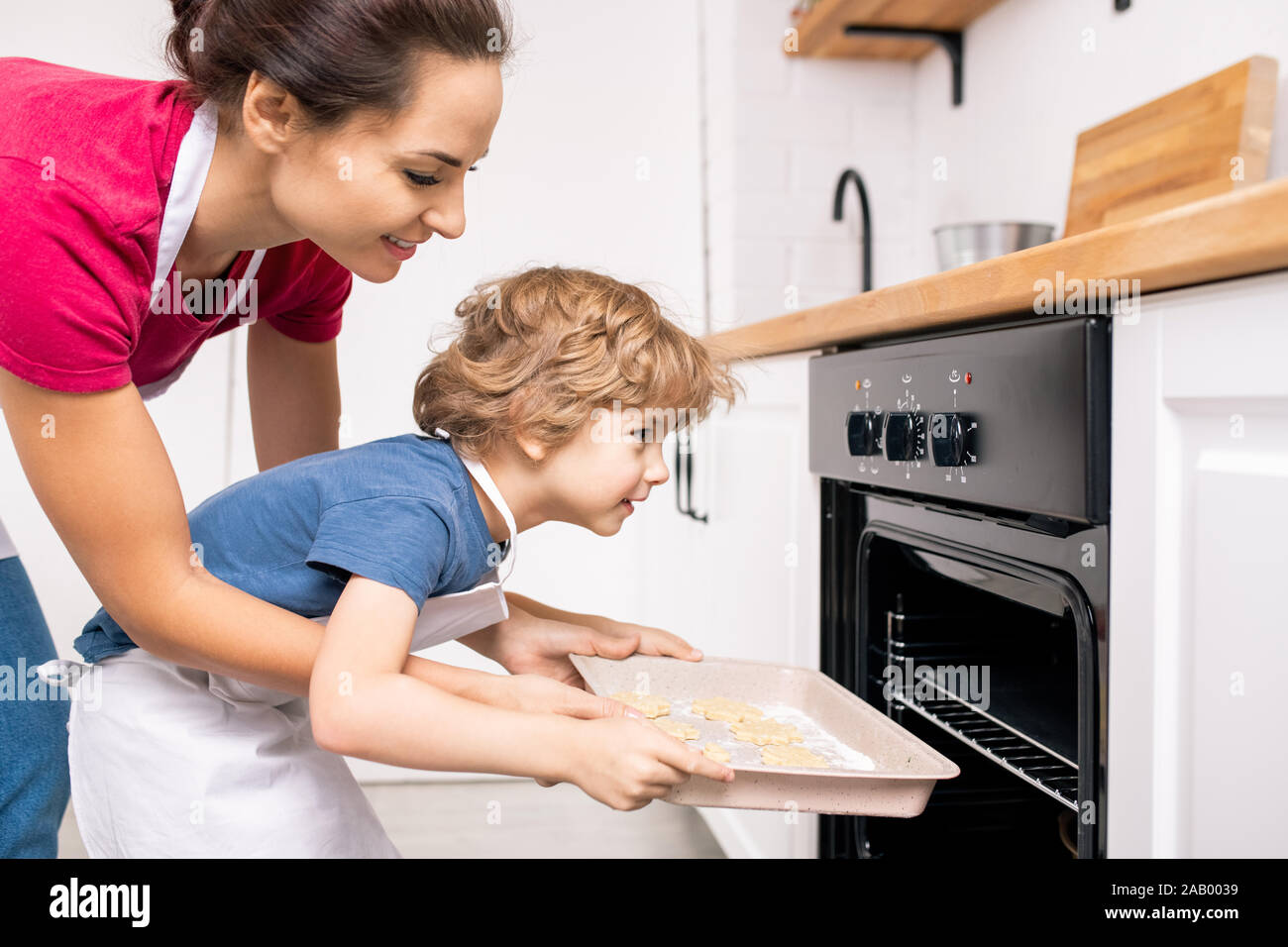 Junge weibliche ihr kleiner Sohn helfen, Fach mit raw-Cookies in offene Backofen während beide biegen nach vorne Stockfoto