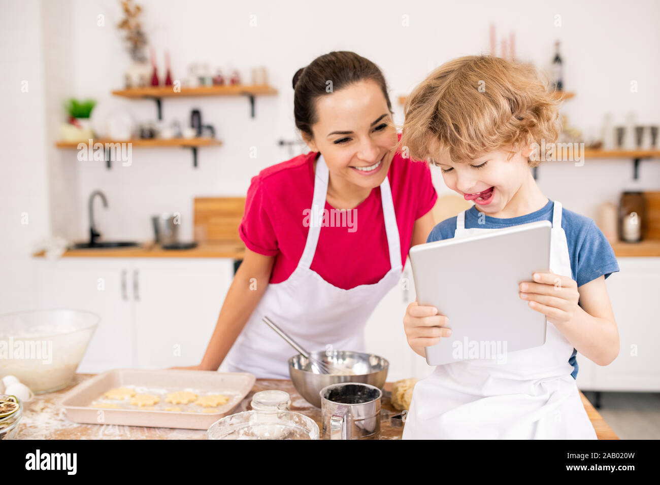 Adorable fröhliches Kind mit Touchpad, seine Mutter video Rezept der etwas wirklich lecker angezeigt, während Sie was zu kochen Stockfoto