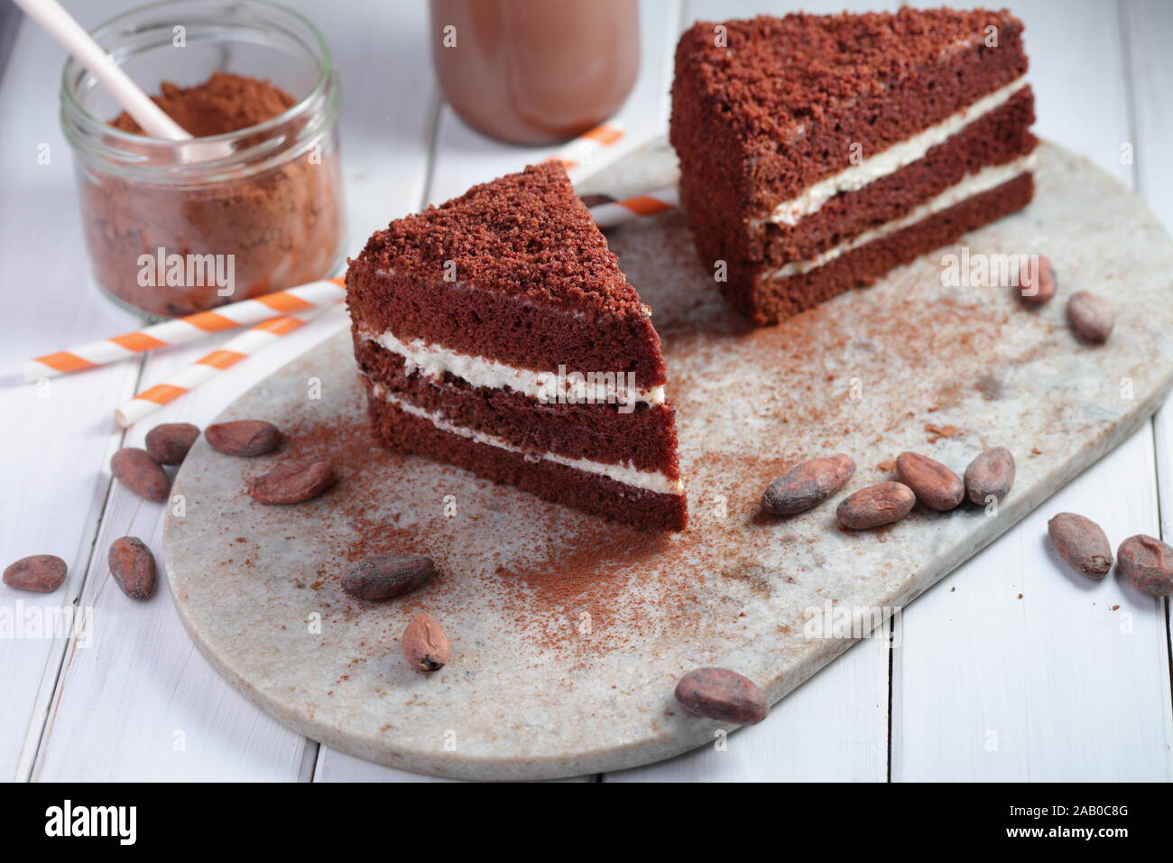 Zwei Scheiben Schokolade Kuchen, heiße Schokolade in einer Flasche, Milchpulver, Kakao und kakaohaltige Bohnen auf einem rustikalen Tisch Stockfoto