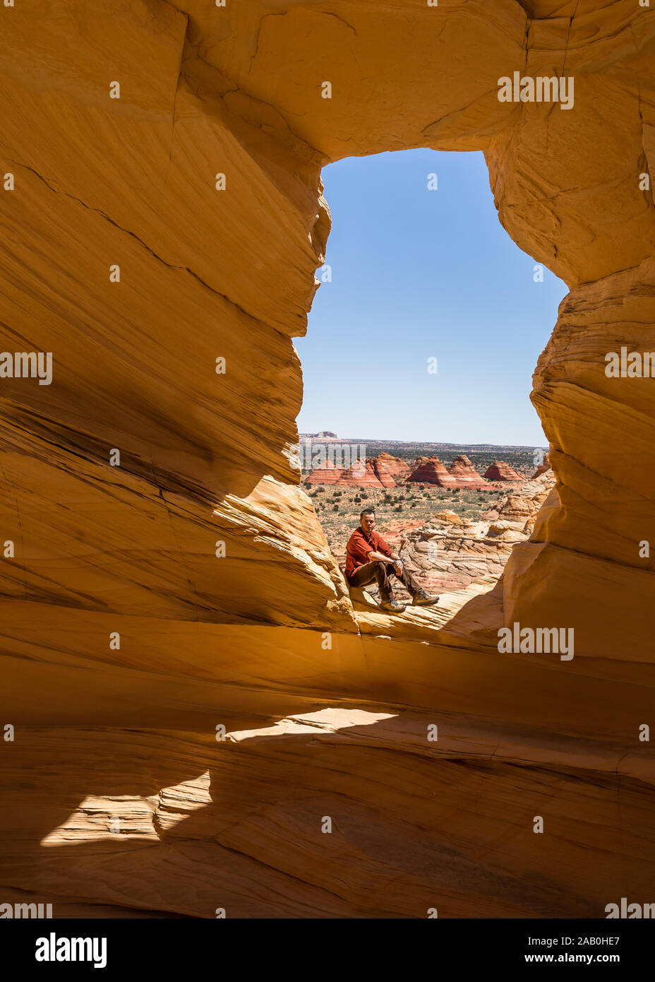 Mann sitzend in die Kamera schaut in einen Torbogen aus der Nische, mit Blick auf Teepee Sandsteinformationen der Coyote Buttes North wilderne Stockfoto