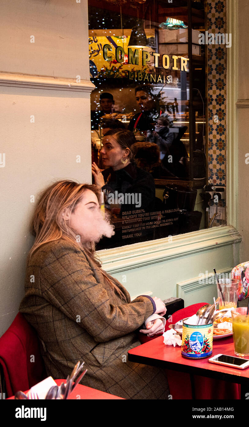 Frau sitzt außerhalb Cafe nach dem Mittagessen ausatmen vaping Rauch. South Kensington, London, England, UK. Stockfoto