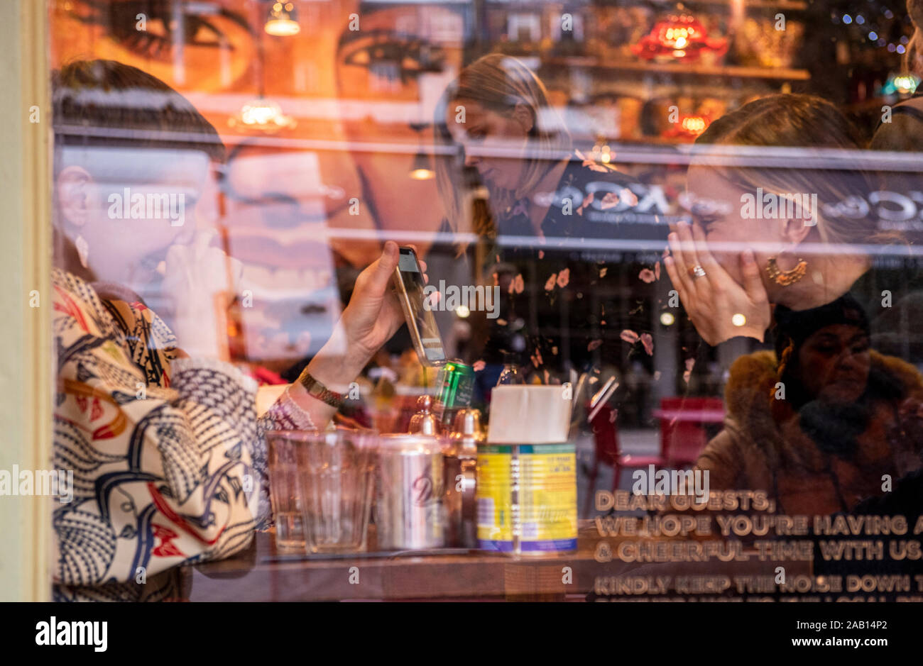Zwei weibliche Freunde essen Mittagessen durch Fenster in Cafe lachend und an Smartphone. South Kensington, London, England, UK. Stockfoto