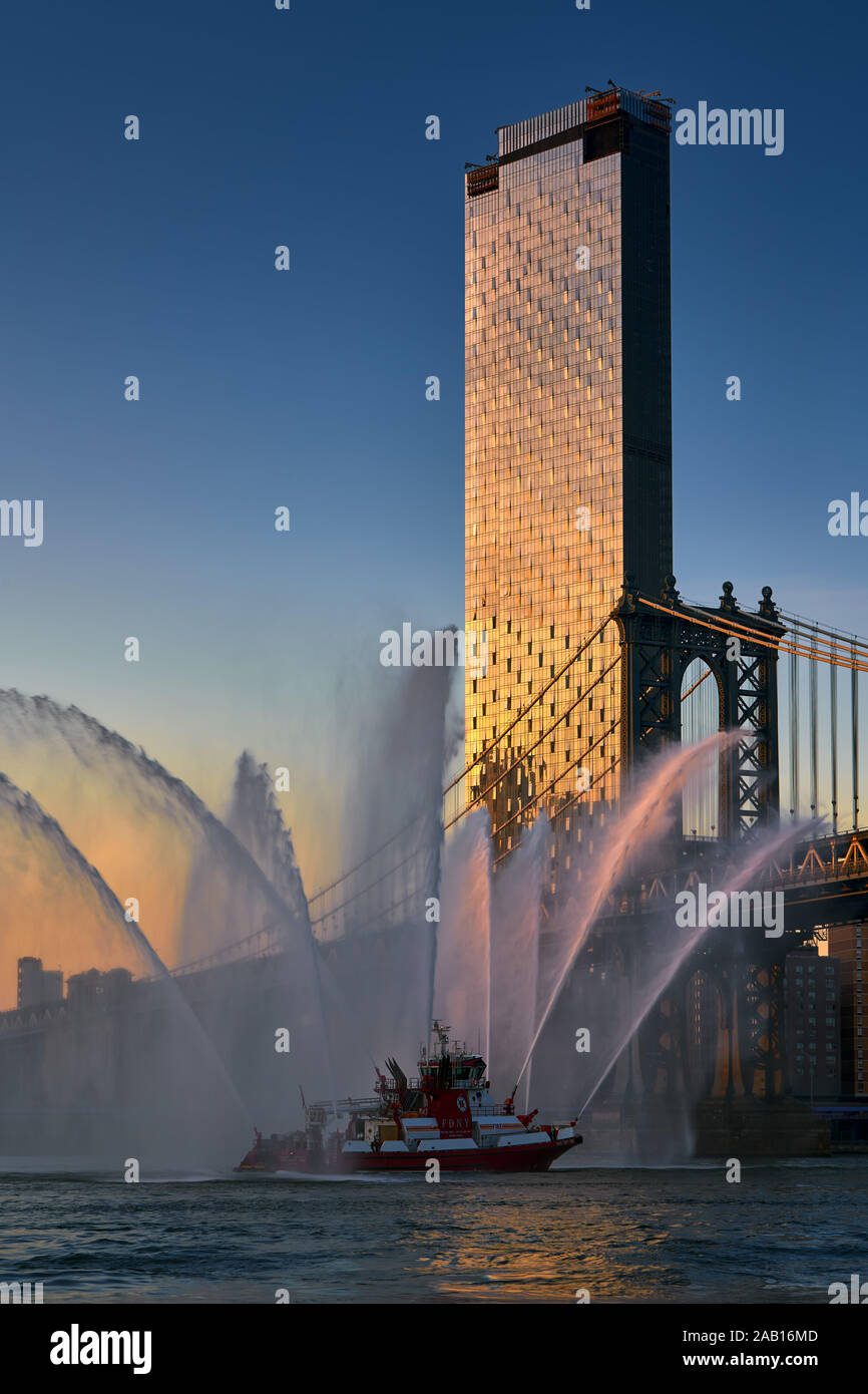 Ein Manhattan Square skyscraper mit der Manhattan Bridge und FDNY feuerlöschboot Wasserstrahlen bei Sonnenuntergang. Lower Manhattan, New York City, NY, USA Stockfoto