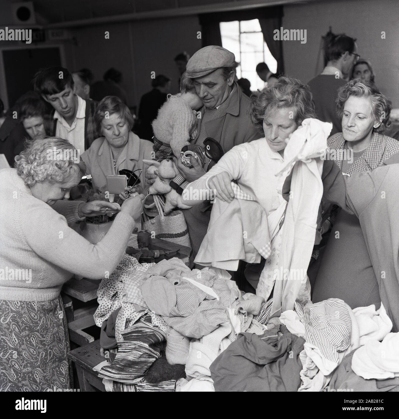 In den 1960er Jahren, historisch, waren die Menschen in einer Dorfhalle bei einem durcheinander gerodeten Verkauf, der durch die Backstein-A-Brac- und Secondhand-Kleidung auf einem Stall, England, Großbritannien, gestapelt wurde. Stockfoto