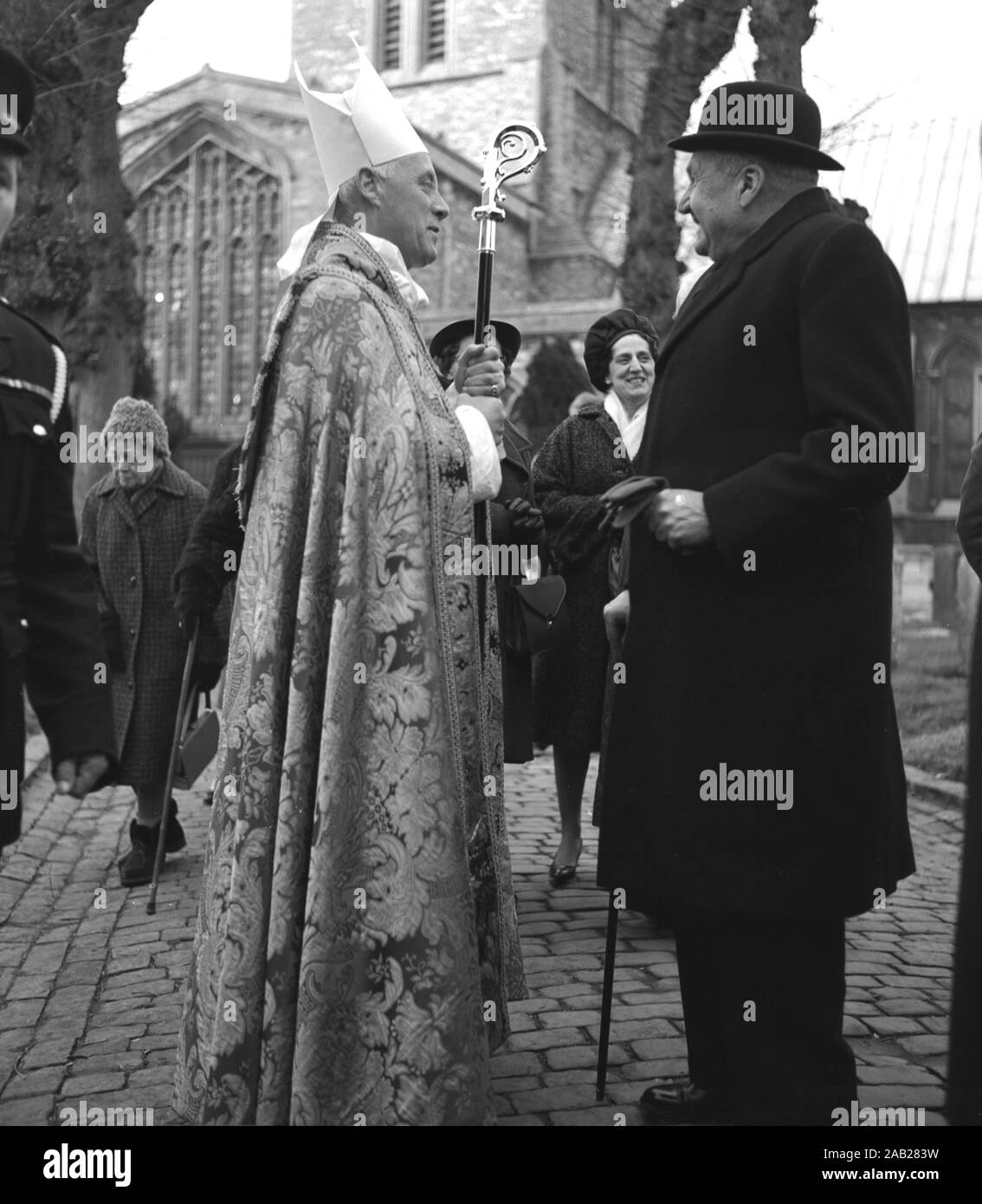 1964, historisch, ein hochrangiger römisch-katholischer Priester, der einen gestickten Dalmatiker oder ein Kleid, eine Pretta (Hut) trägt und einen Crosier oder einen Stab hält, der mit einem älteren Herrn spricht, der einen Melonenhut vor der St Mary's Church, Aylesbury, England, Großbritannien, trägt. Stockfoto