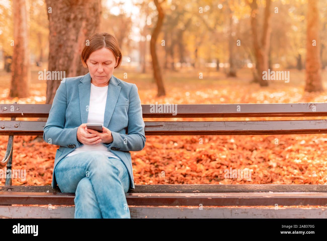 Frau mit Mobiltelefon auf Pausen während der Arbeitszeit beim Sitzen auf einer Parkbank an sonnigen Herbsttag Stockfoto
